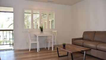 A living room with a brown leather sofa, a wooden coffee table, a white dining table with two chairs, and a window with blinds.