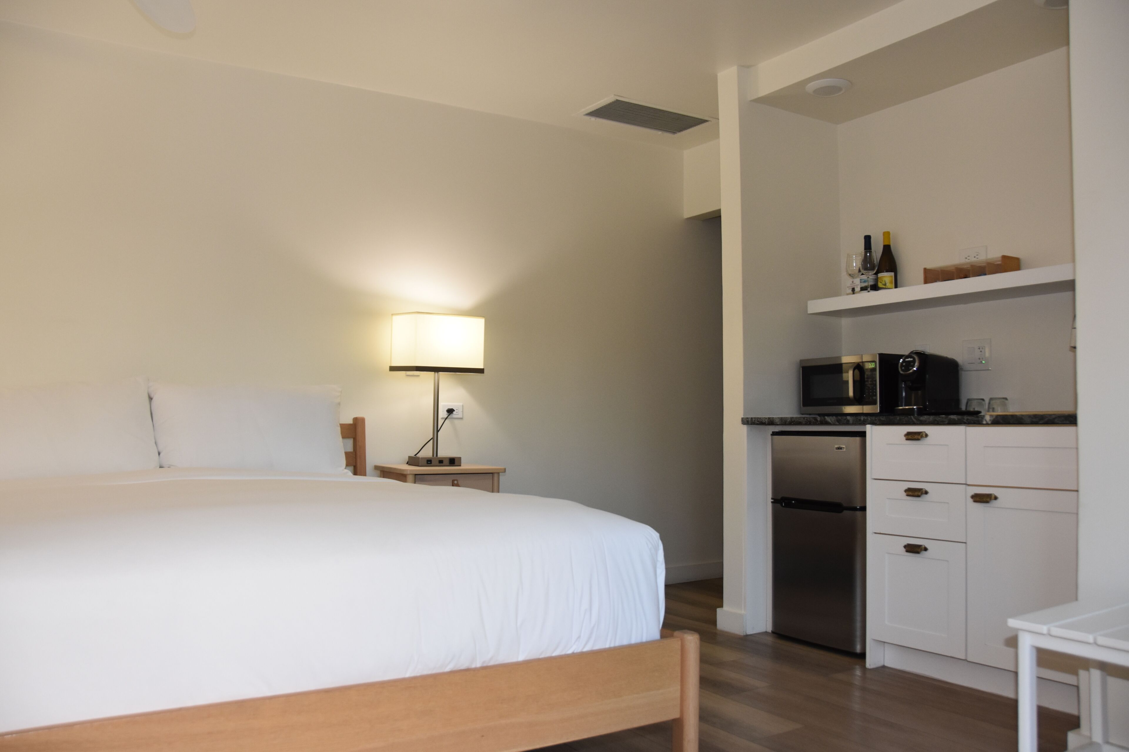 Une chambre comprenant un lit, une table de chevet, une lampe et une kitchenette équipée d’un four à micro-ondes, d’un réfrigérateur et d’étagères.