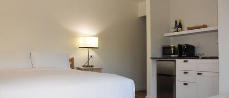 A bedroom with a bed, bedside table, lamp, and a kitchenette with a microwave, refrigerator, and shelves.