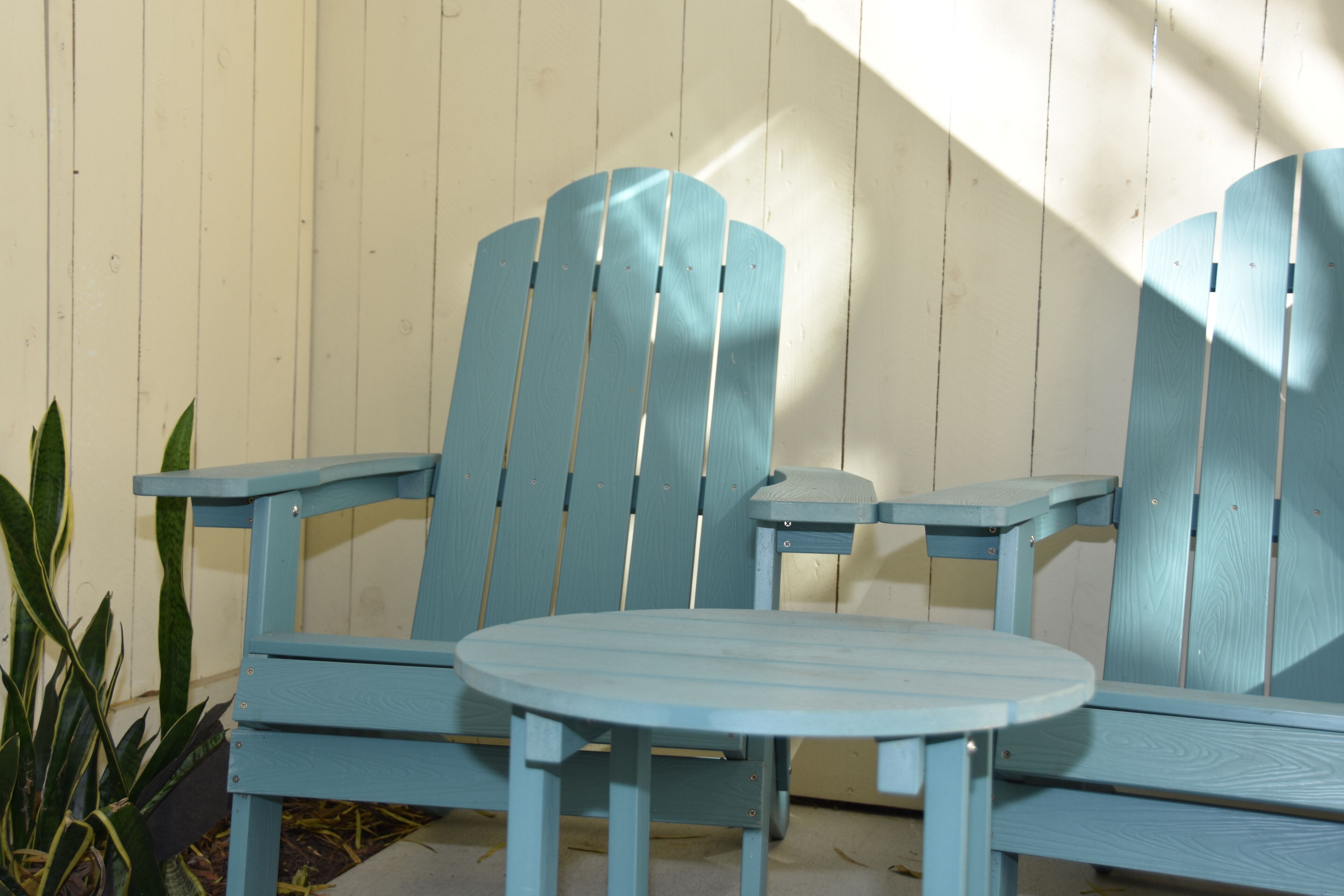 Un ensemble de chaises Adirondack bleues et une petite table ronde, probablement destinées à être utilisées à l’extérieur pour les repas, est placé contre un mur en bois, avec une plante sur le côté gauche.