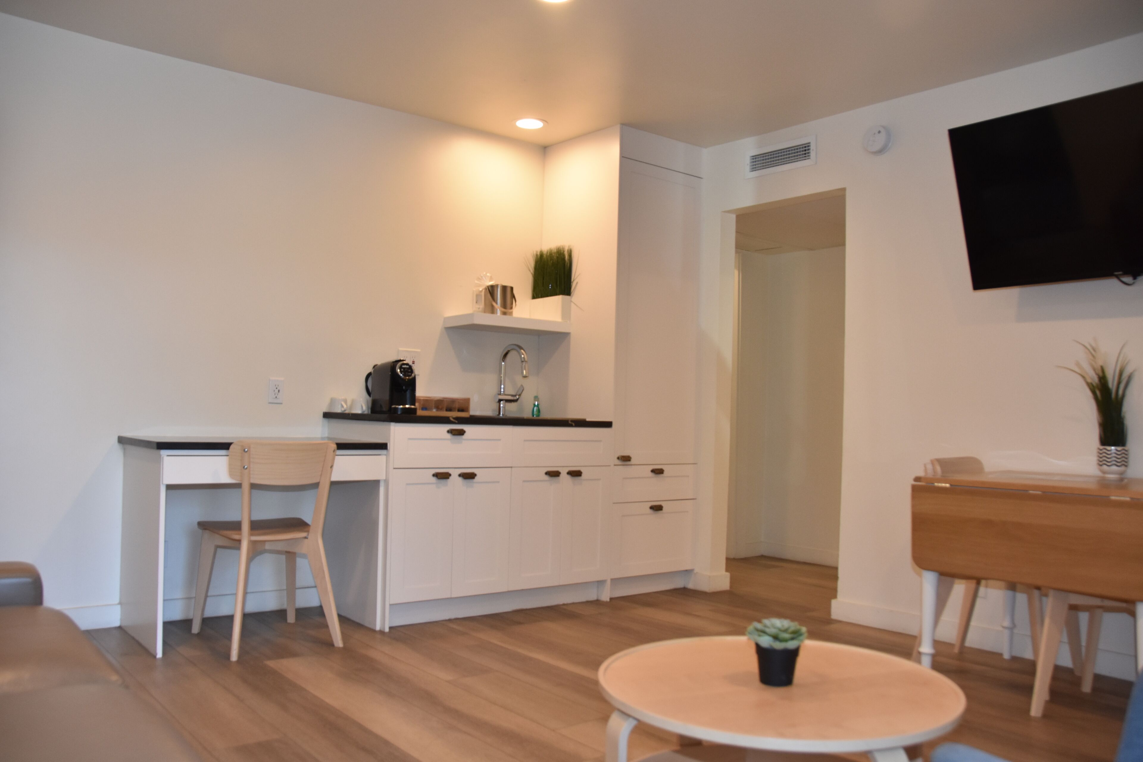 A modern kitchen with white cabinets, a wooden dining table, and a TV mounted on the wall.