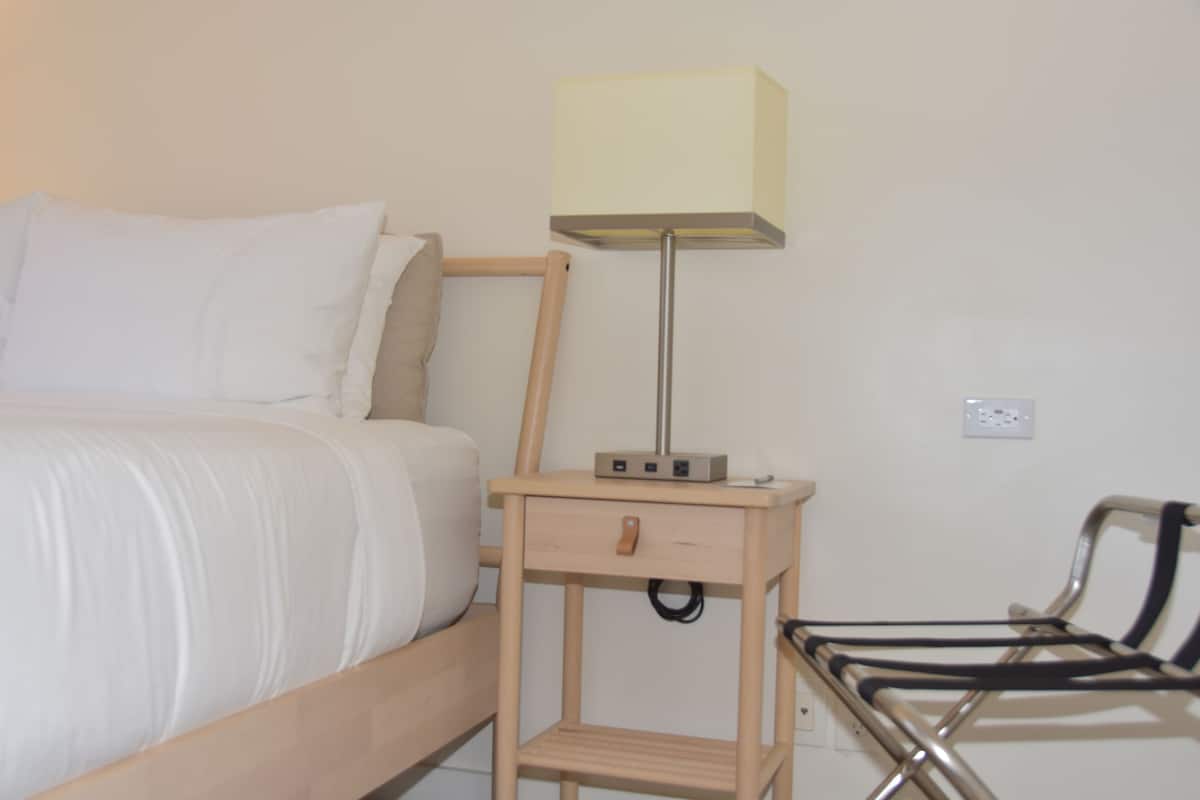 A neatly made bed with white linens, a bedside table with a lamp, and a chair with a metal frame.