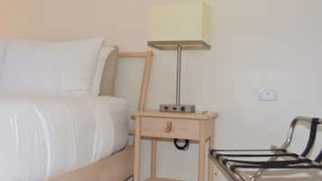 Una cama bien hecha con sábanas blancas, una mesita de noche con lámpara y una silla con estructura metálica.