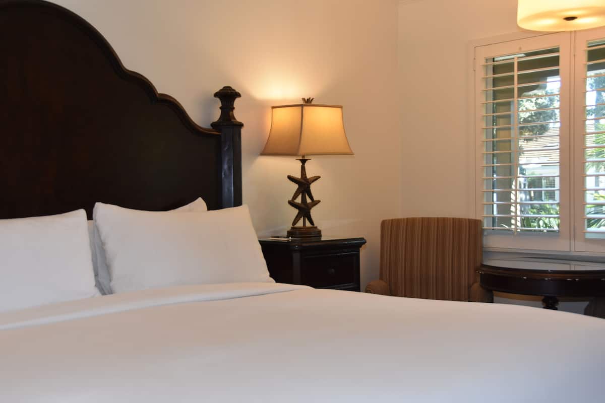 A hotel room with a large bed, a nightstand with a lamp, a chair, and a window with blinds.