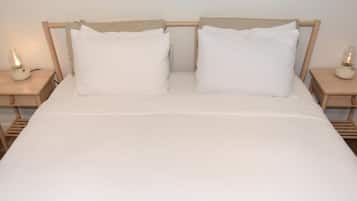 Uma cama com roupa de cama branca, duas almofadas e cabeceira de madeira. Há duas mesas de cabeceira com luminárias de cada lado da cama.