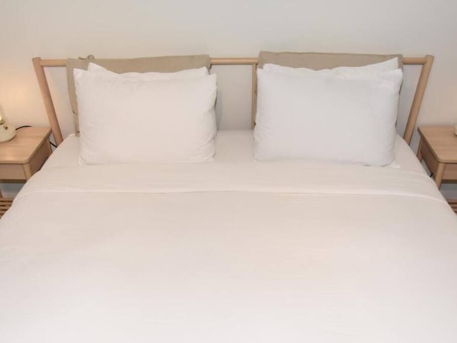 Uma cama com roupa de cama branca, duas almofadas e cabeceira de madeira. Há duas mesas de cabeceira com luminárias de cada lado da cama.