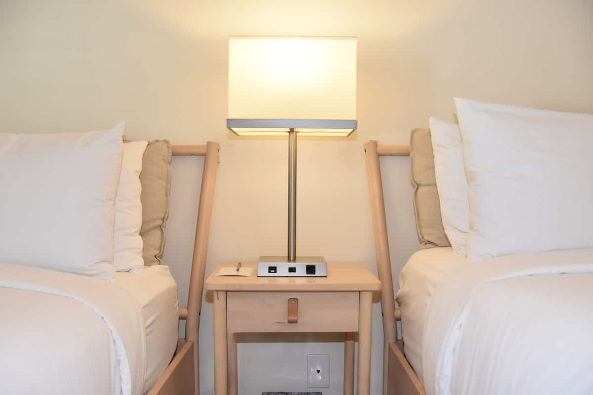 A hotel room with two beds, a bedside table with a lamp, and a power outlet.