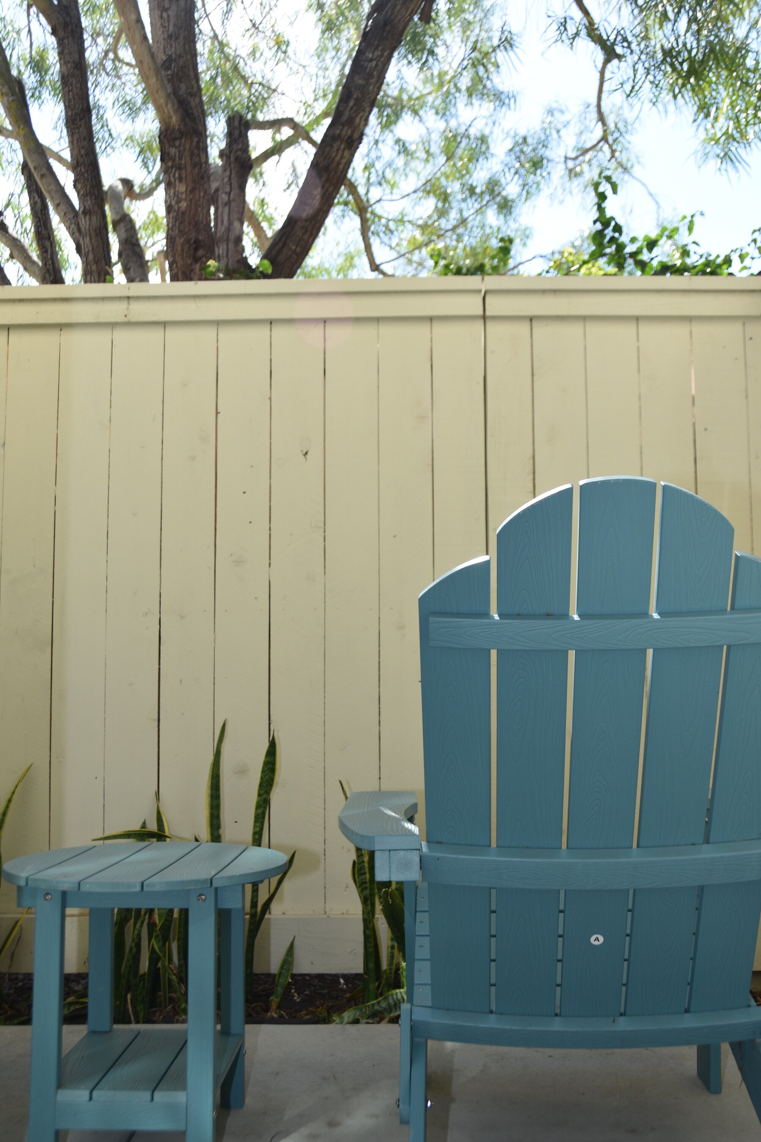 A blue Adirondack chair and a small table in an outdoor setting with a wooden fence and trees in the background.