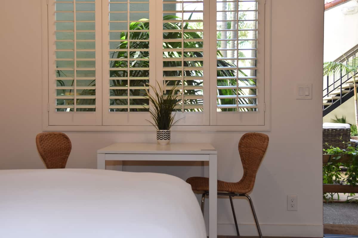 A room with a bed, a small table, two wicker chairs, and a window with blinds.