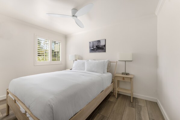 Quarto com cama, ventilador de teto, mesa de cabeceira com abajur, janela com persianas e quadro na parede.