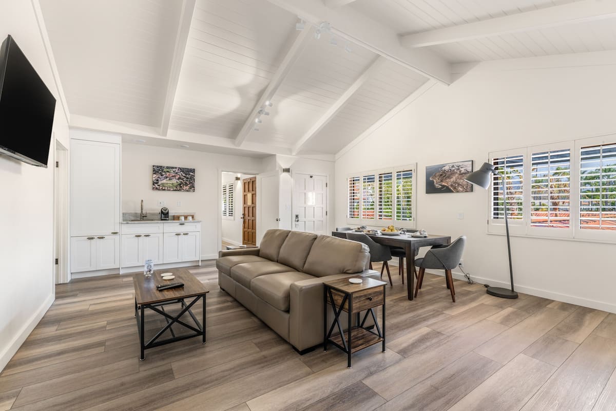 A spacious living area with a high ceiling, a sofa, a coffee table, and a dining table with chairs. There is a kitchen in the background with white cabinets and a television mounted on the wall.
