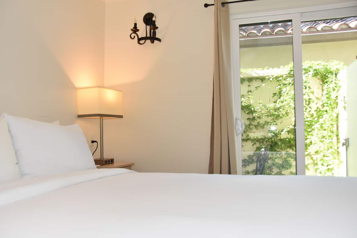 A hotel room with a bed, a bedside lamp, a wall-mounted light fixture, and a view of greenery through a window.