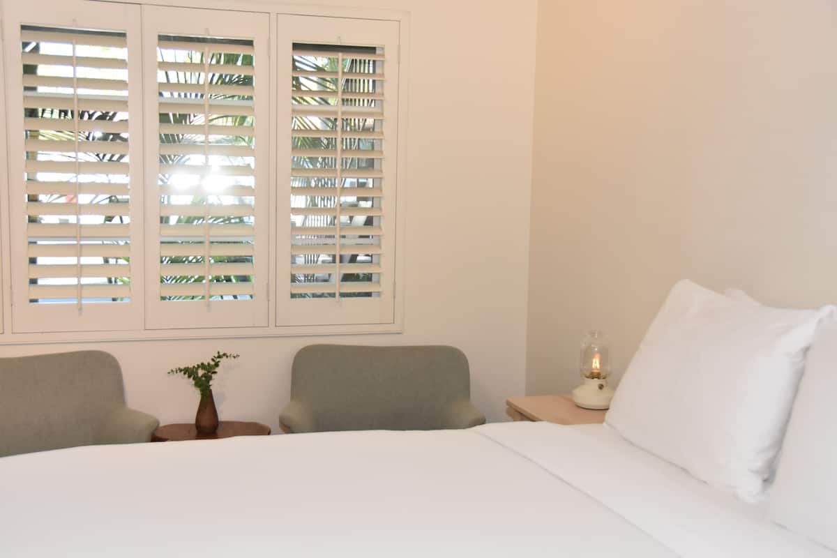 A bedroom with a bed, two chairs, a nightstand with a lamp, and a window with blinds.