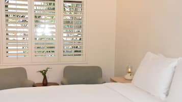 Un dormitorio con cama, dos sillas, una mesita de noche con lámpara y ventana con persianas.
