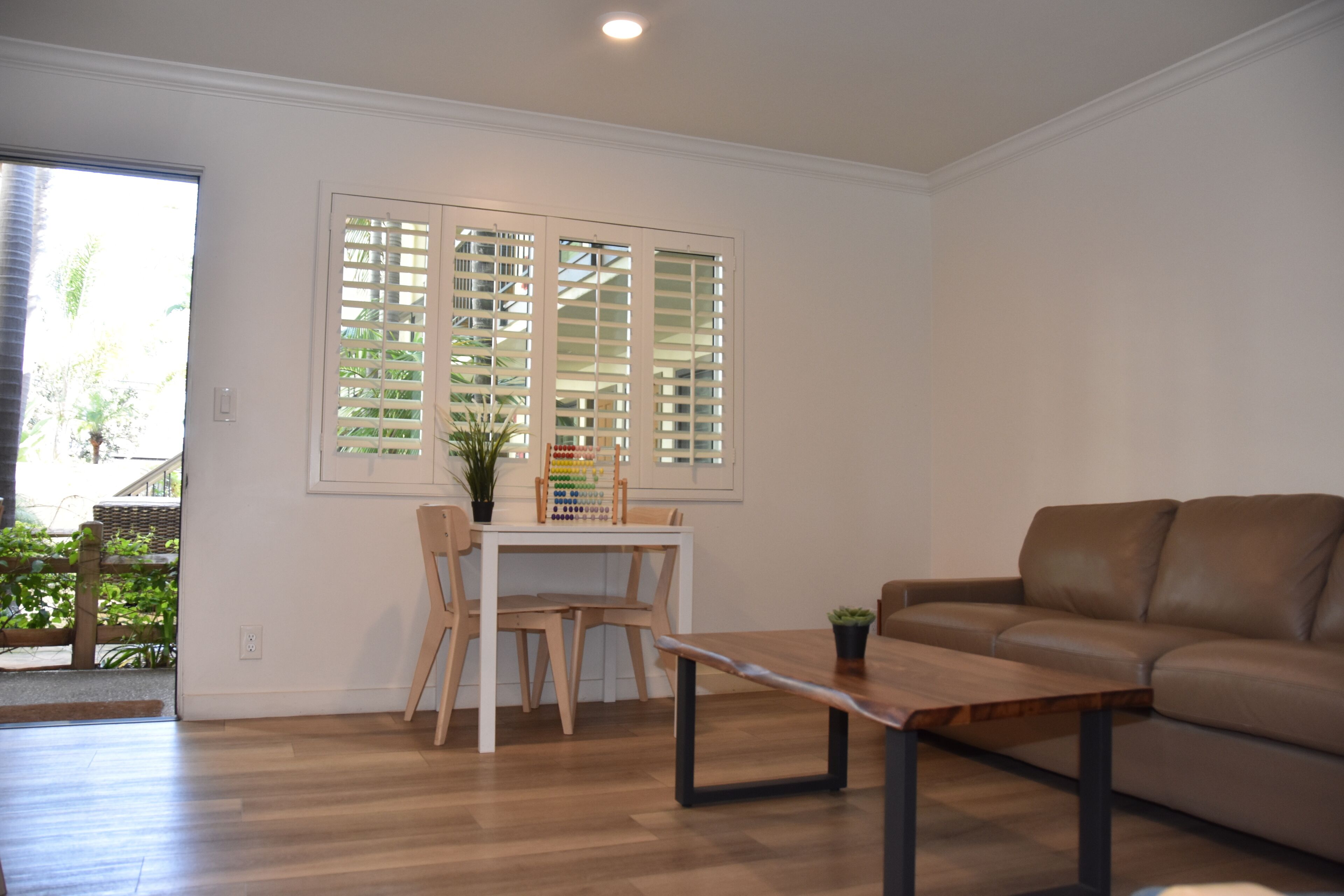 A living room with a brown leather sofa, a wooden coffee table, and a dining table with chairs. There is a window with blinds and a potted plant on the table.