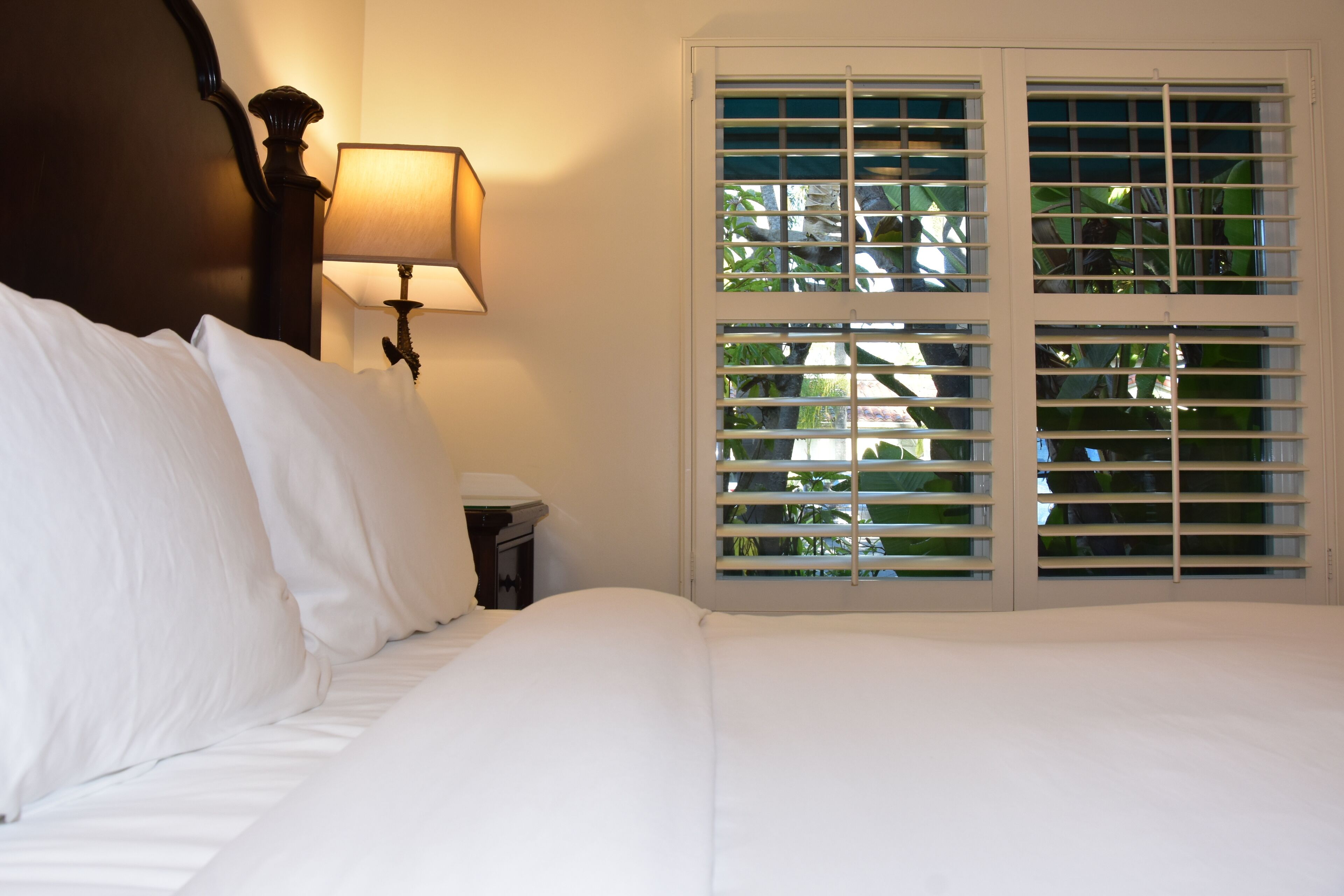 A neatly made bed with white linens, a wall-mounted lamp, and a window with blinds.