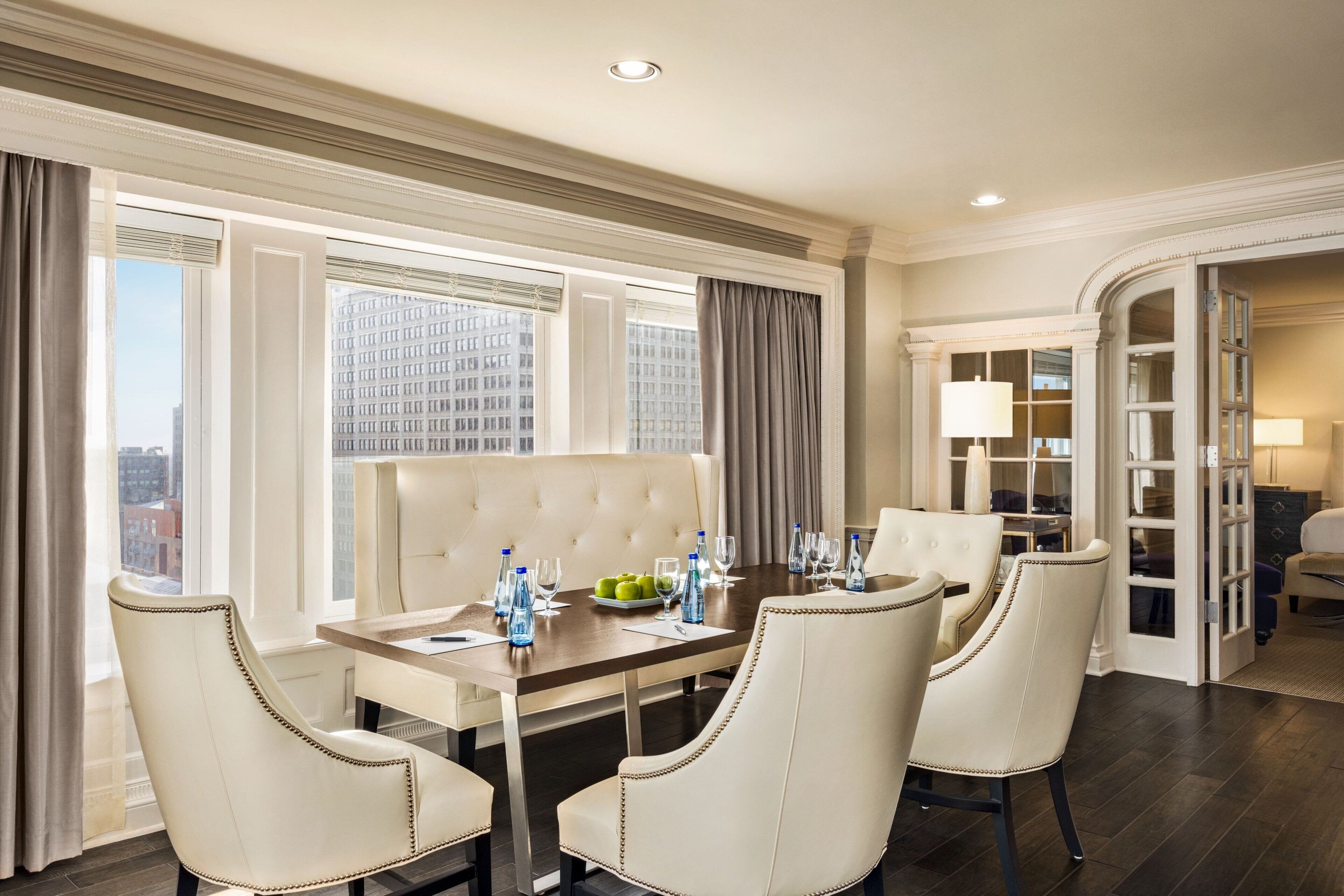 A hotel dining room with a large table, four chairs, and a view of the cityscape through large windows.