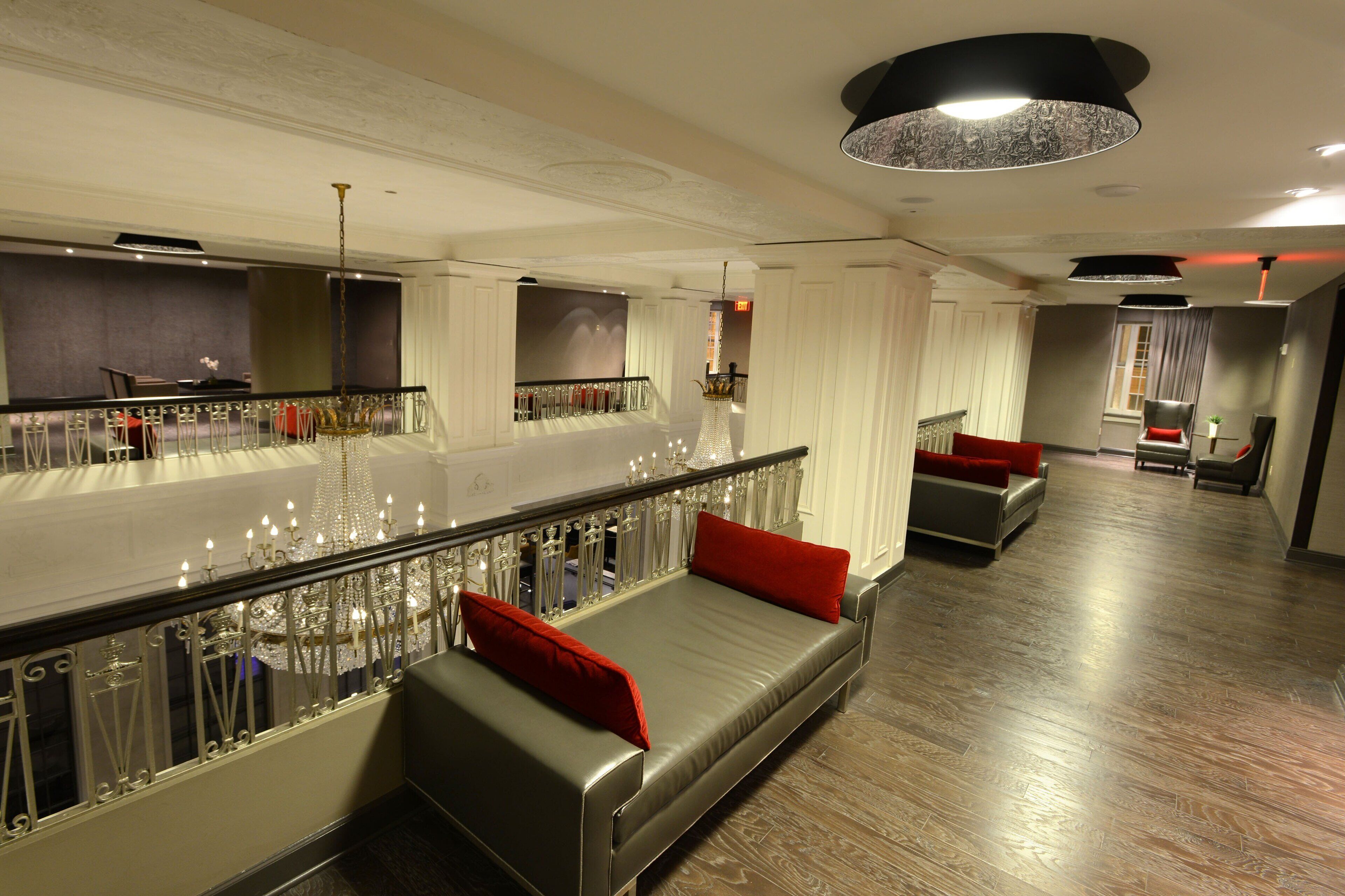 A modern hotel lobby with a long reception desk, a seating area with a sofa and red cushions, and a staircase with a decorative railing.