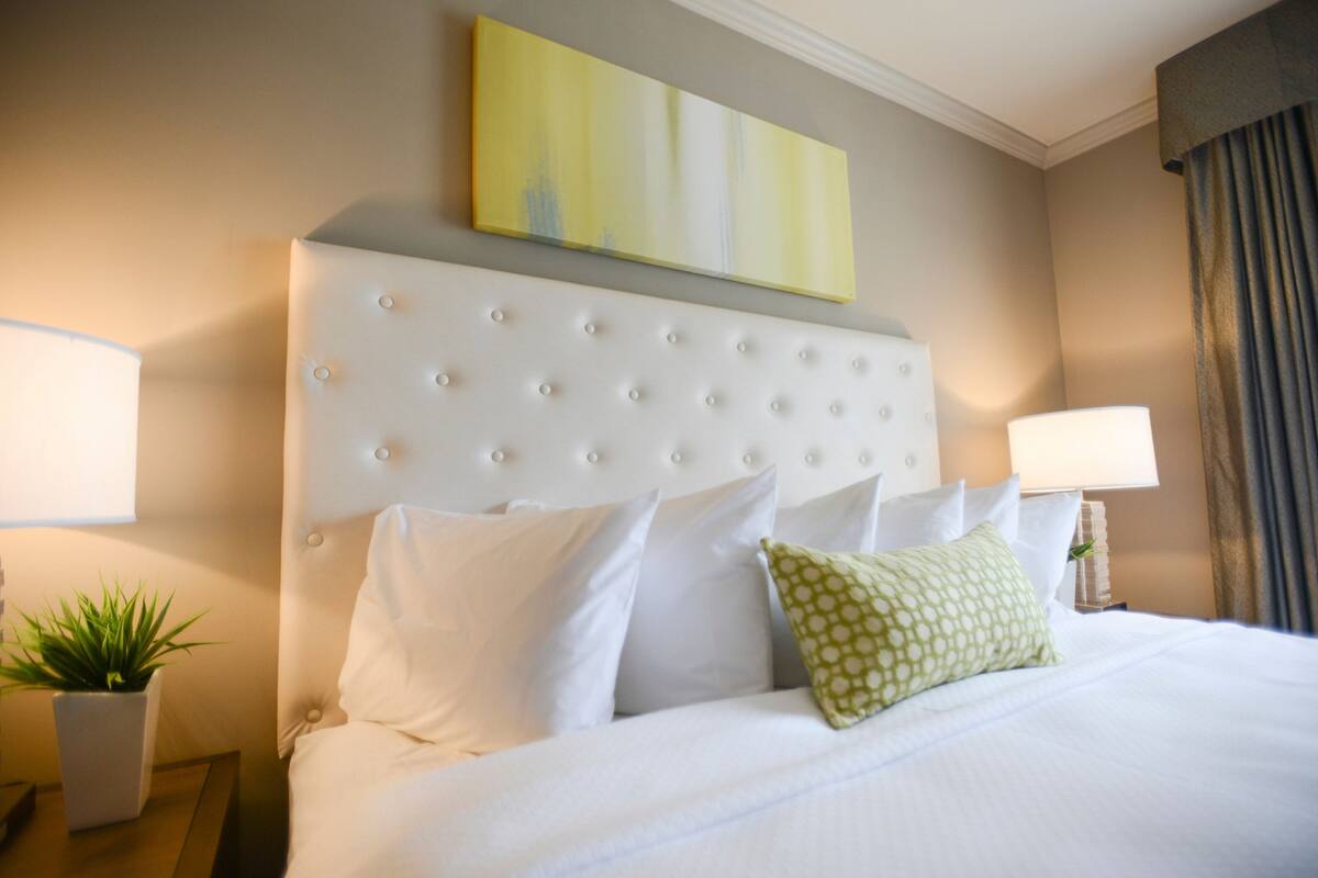 A hotel room with a tufted headboard, white bedding, a green patterned pillow, and a painting on the wall.