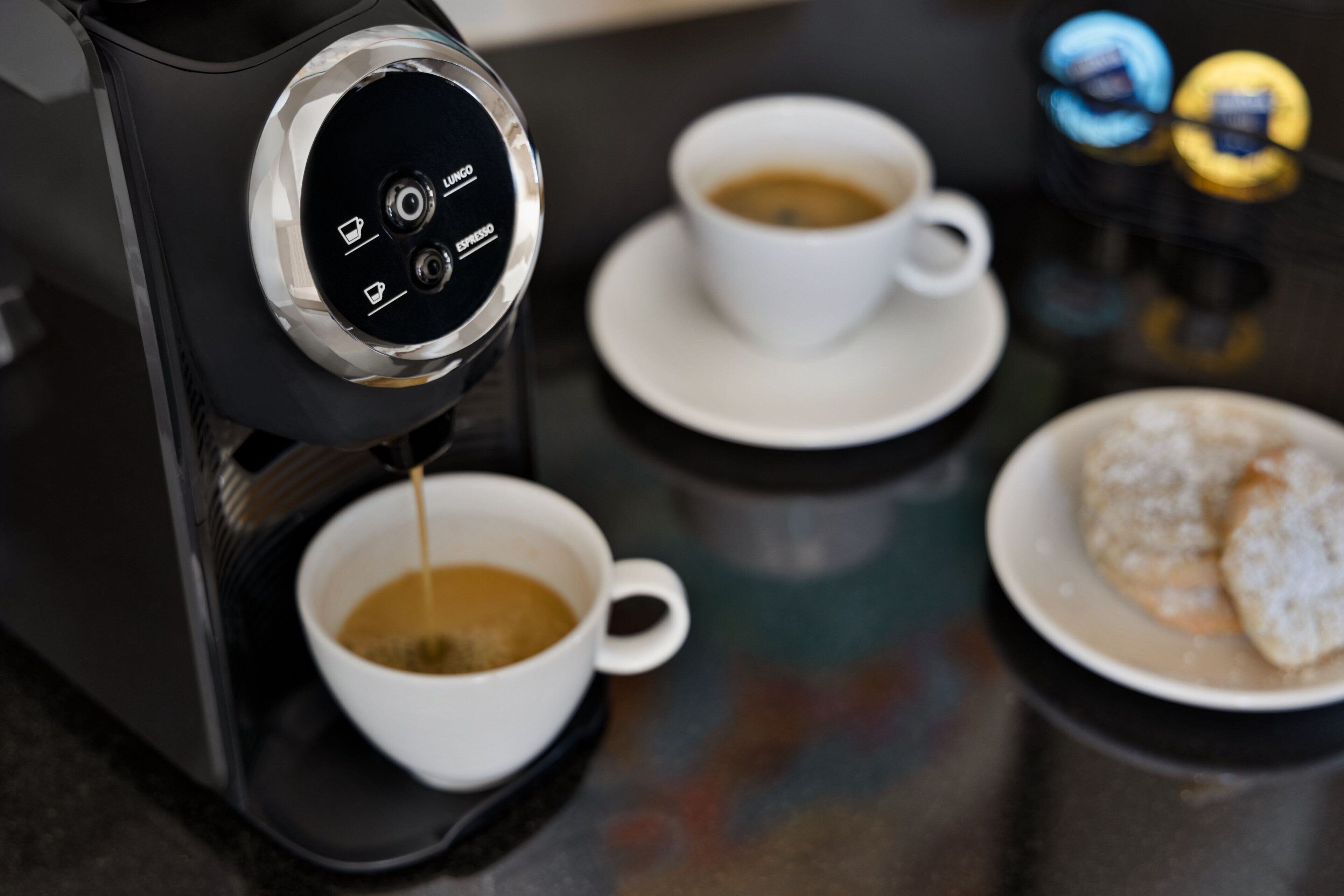 Una cafetera sirviendo café en dos tazas, un plato con galletas y un envase de leche condensada.