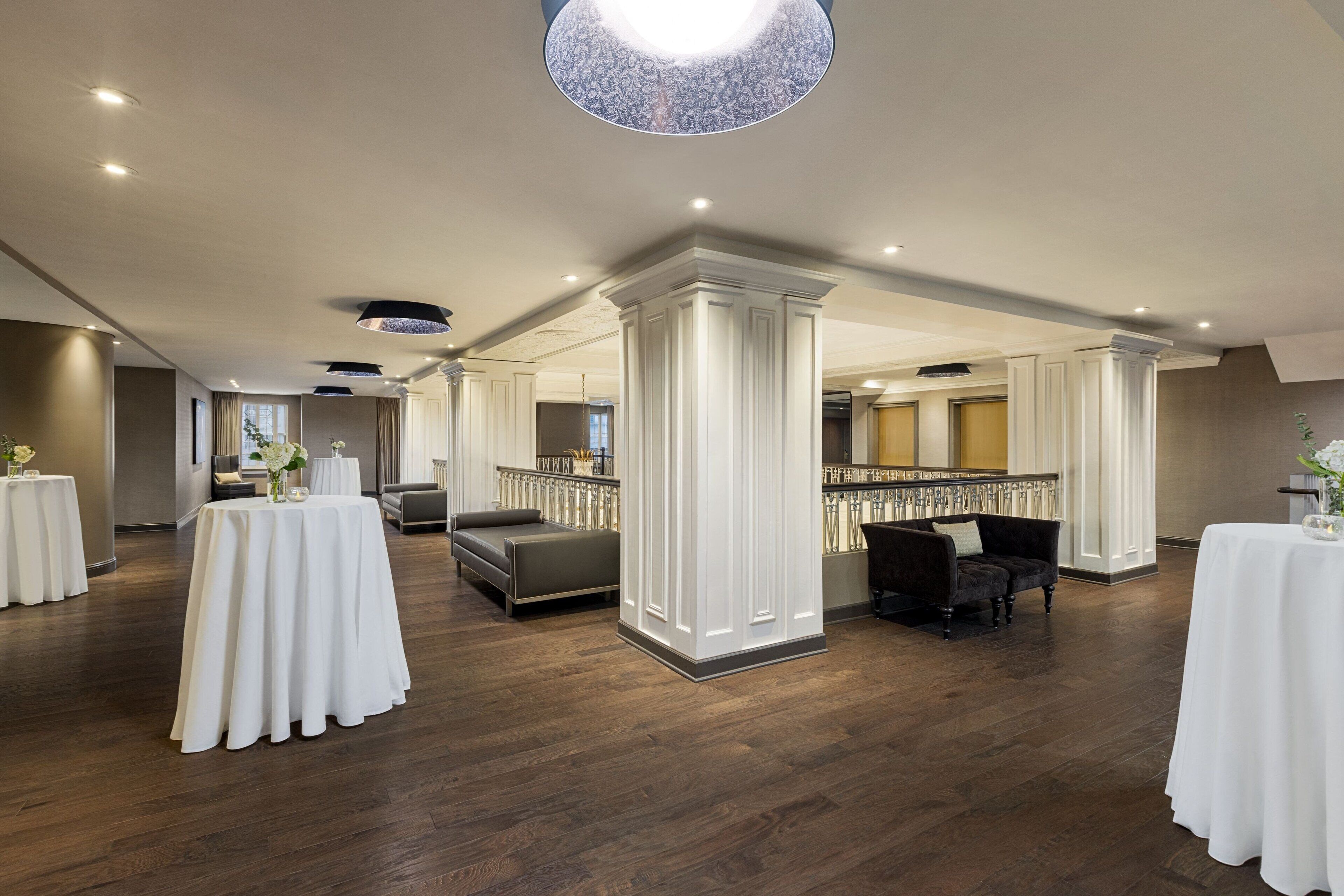A spacious hotel lobby with high ceilings, elegant columns, and a grand staircase. Tables and seating are arranged for guests.
