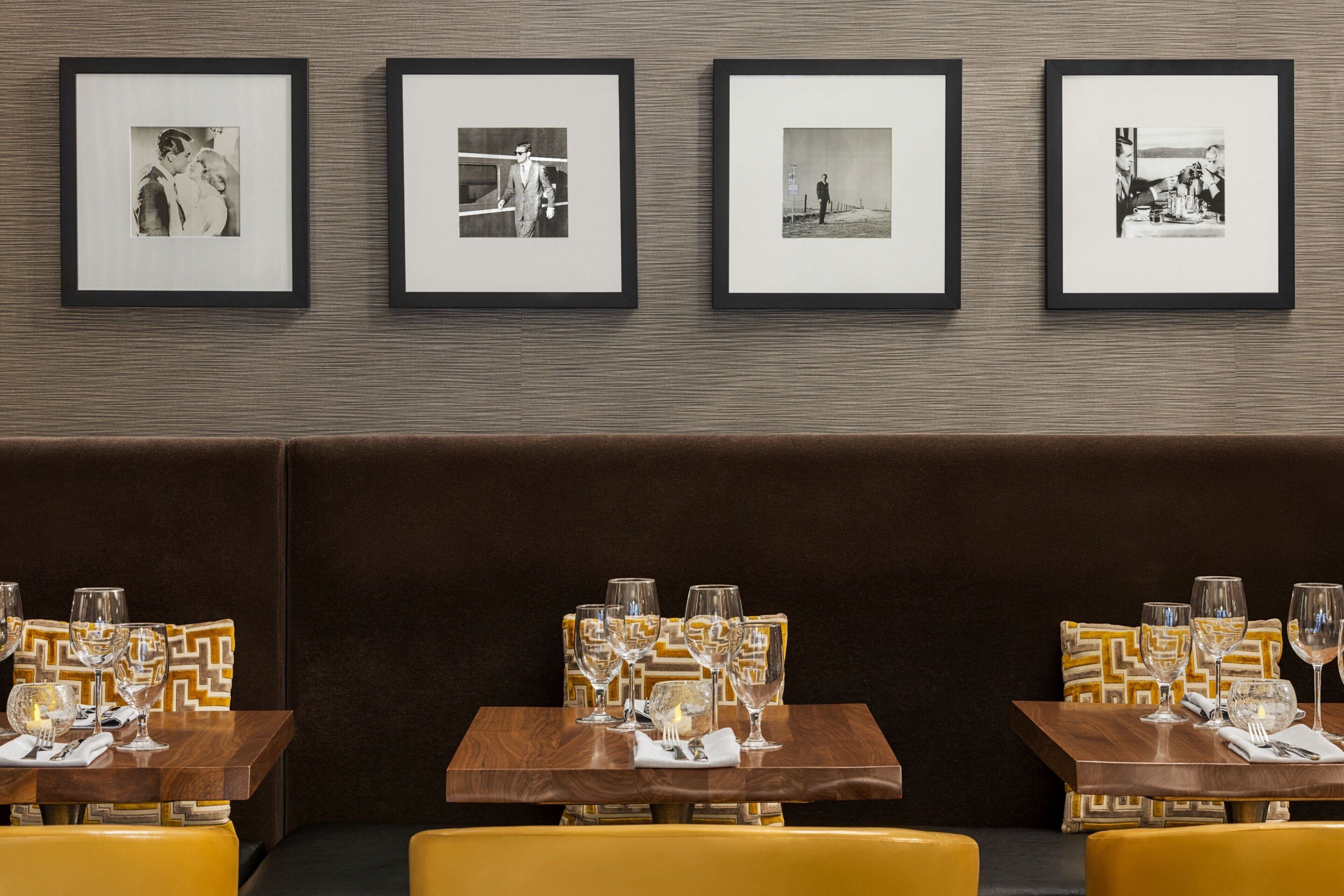 A restaurant with framed black and white photographs on the wall, tables set with wine glasses, and yellow upholstered booths.