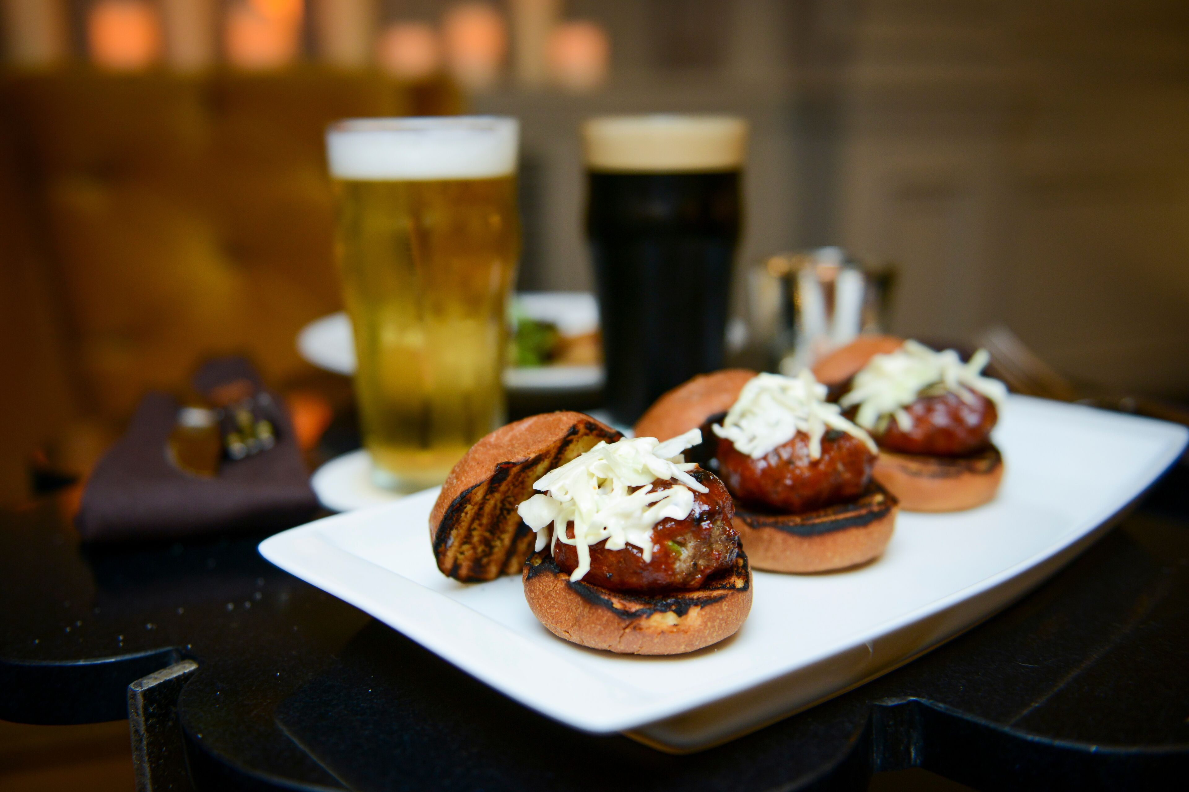 A plate of grilled meatballs with cheese on a white plate, accompanied by a glass of beer and a dark beer in the background.