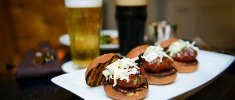 A plate of grilled meatballs with cheese on a white plate, accompanied by a glass of beer and a dark beer in the background.