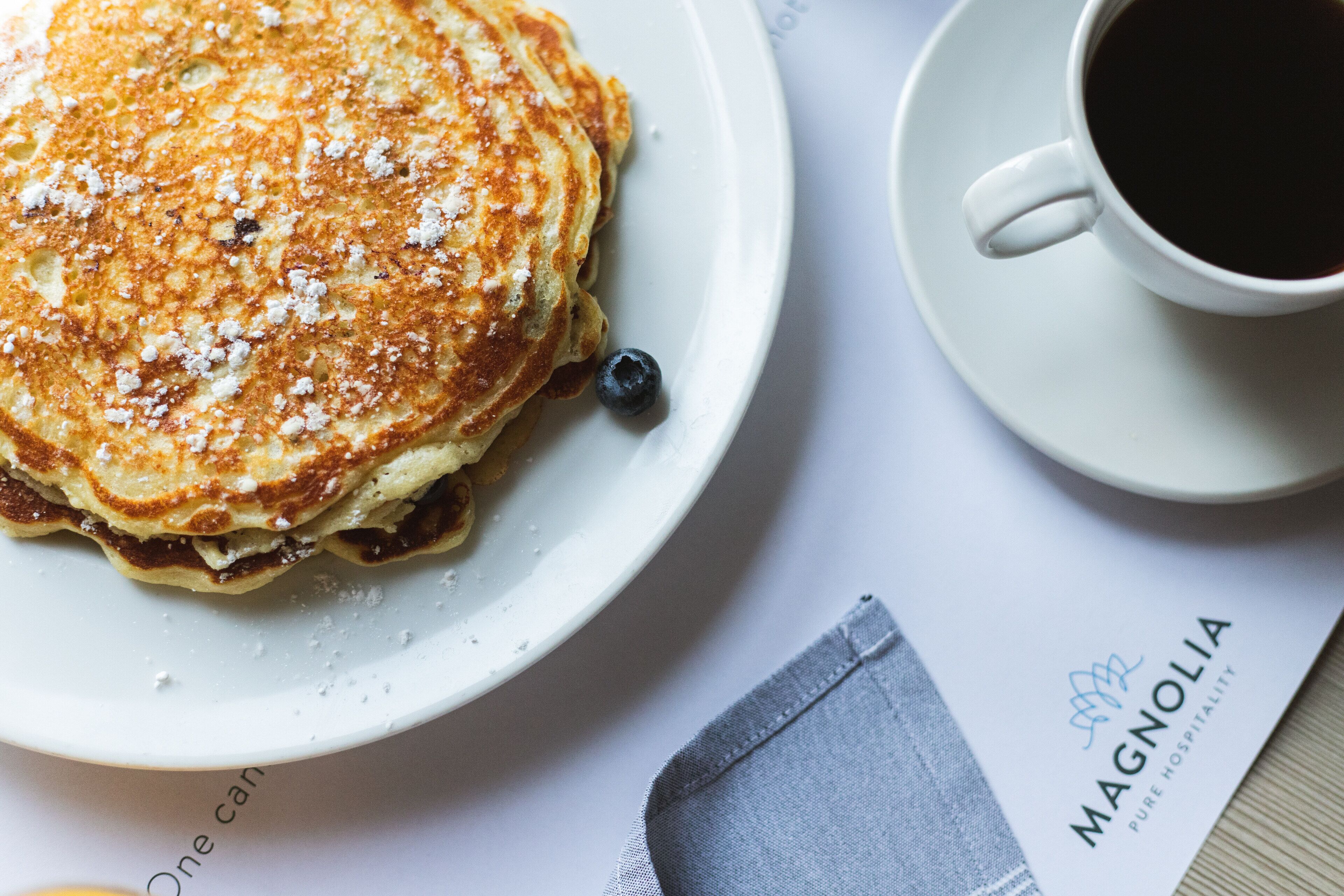 A plate of pancakes with blueberries and powdered sugar, a cup of black coffee, and a napkin with a logo.