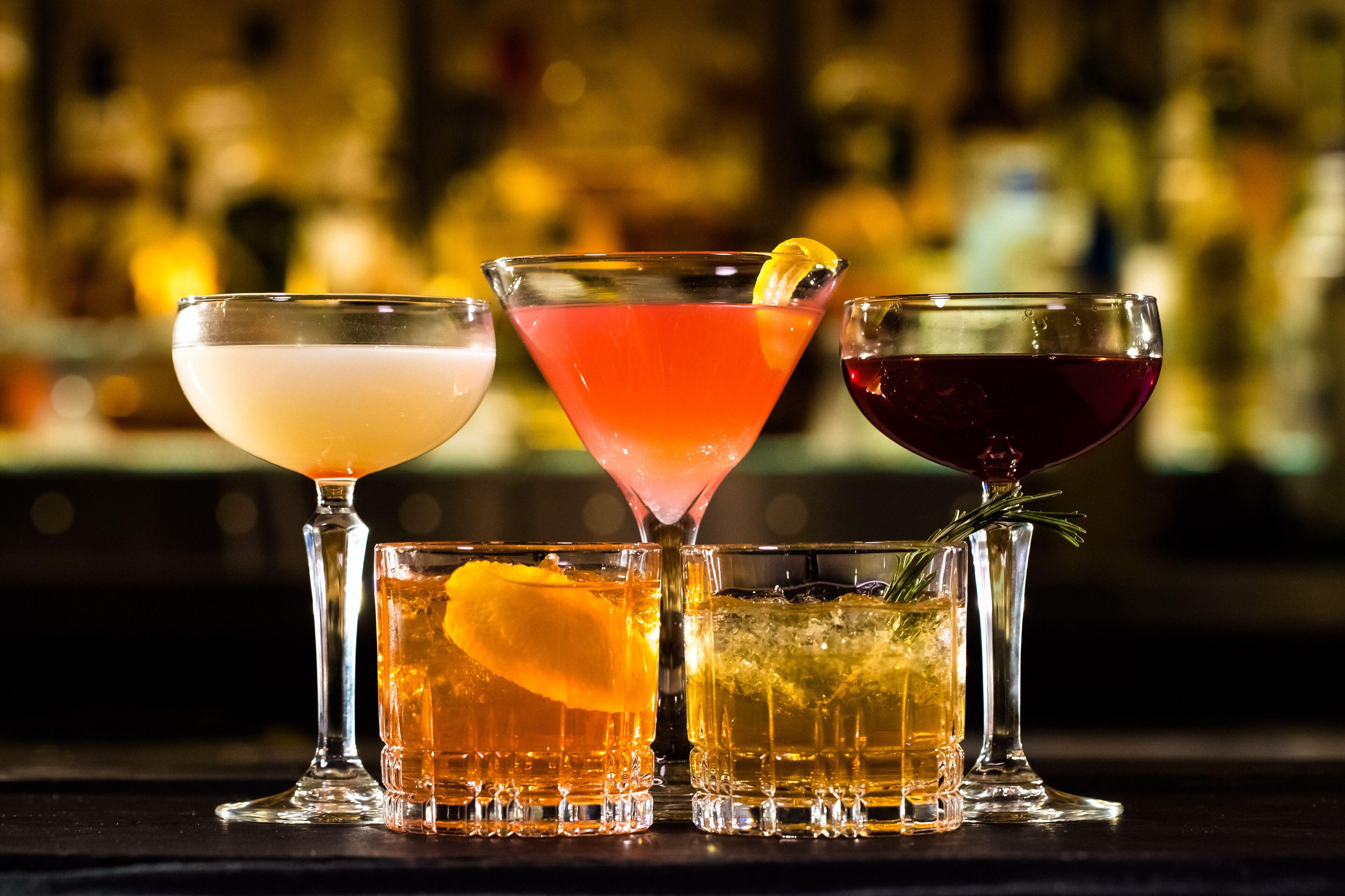 Three different cocktails in various glasses, with a blurred background of bottles.