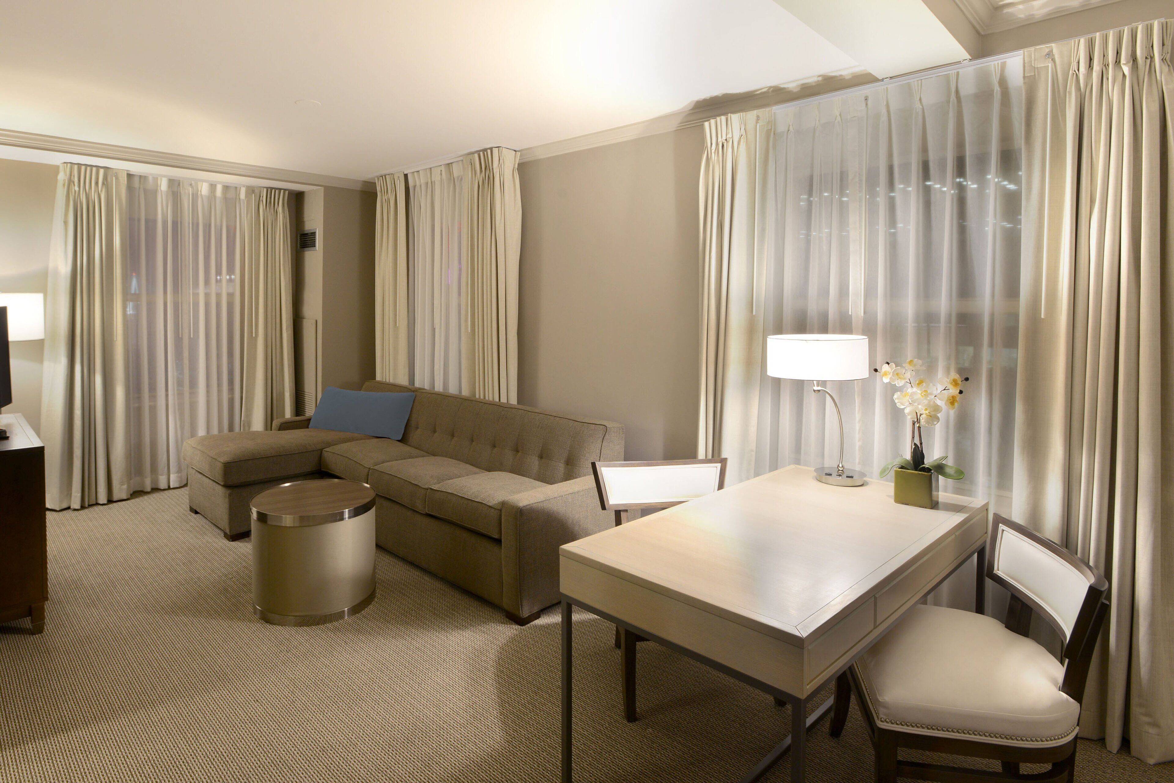 A hotel room with a sofa, a dining table, a chair, a lamp, and a vase with flowers.