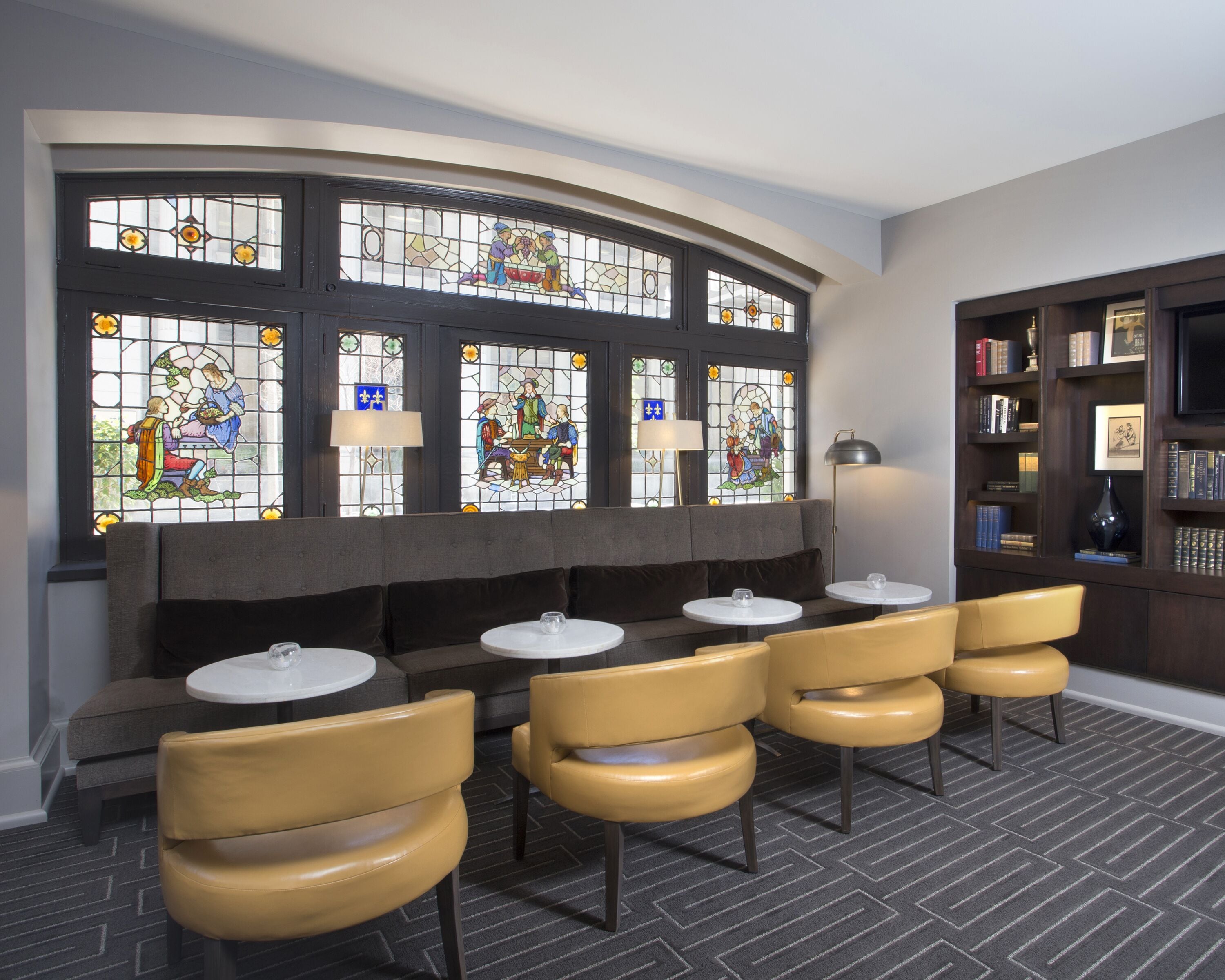 A seating area with a curved bench and round tables, a bookshelf with books and framed pictures, and stained glass windows depicting historical scenes.