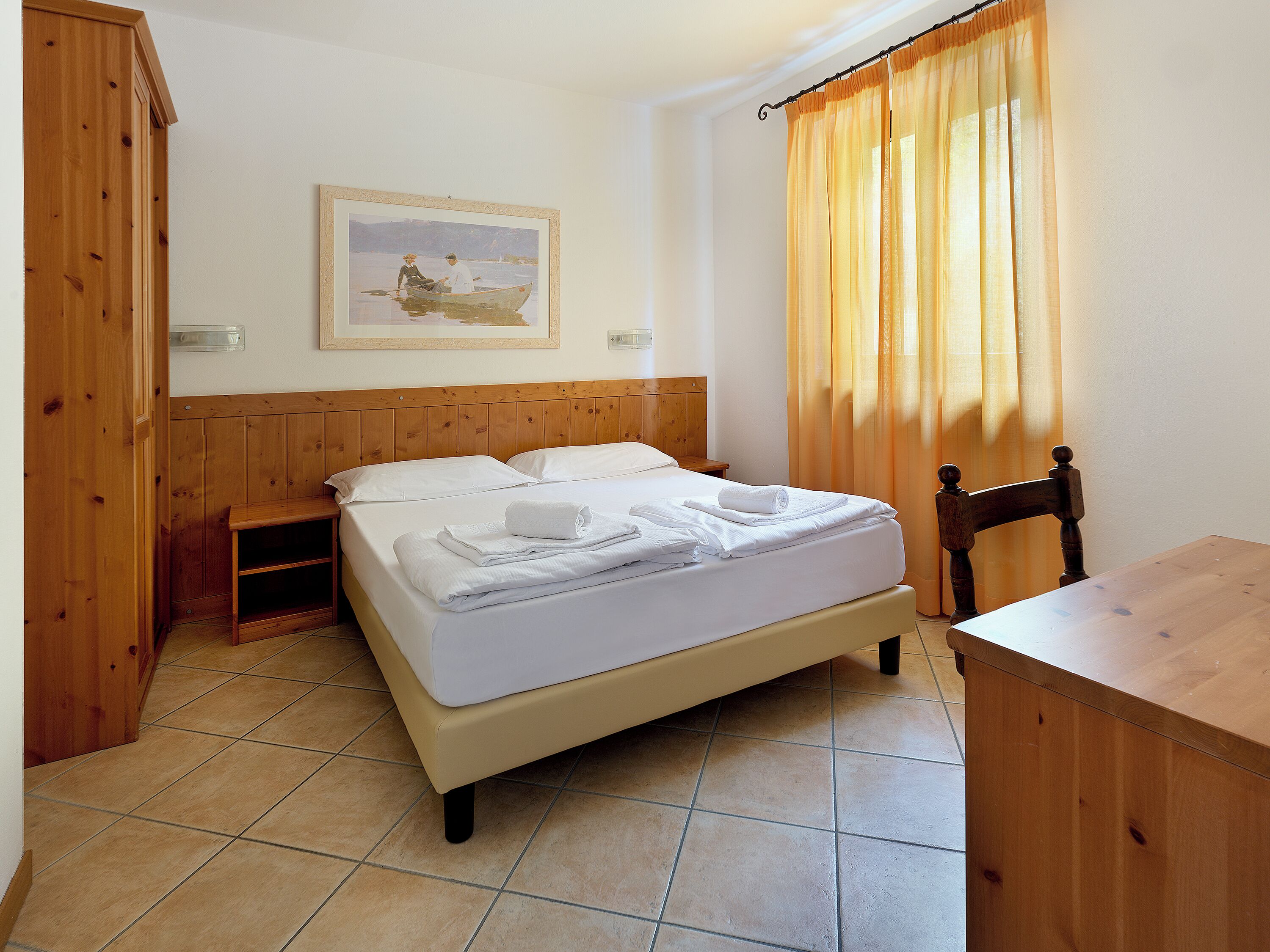 A bedroom with a bed, a wooden headboard, a nightstand, a chair, a table, and a painting on the wall.