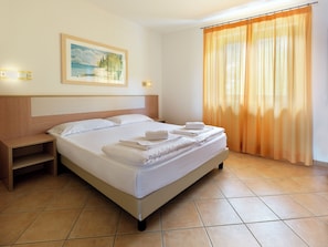 A neatly made bed with white linens and towels, a wooden headboard, a nightstand, a painting on the wall, and a window with orange curtains.