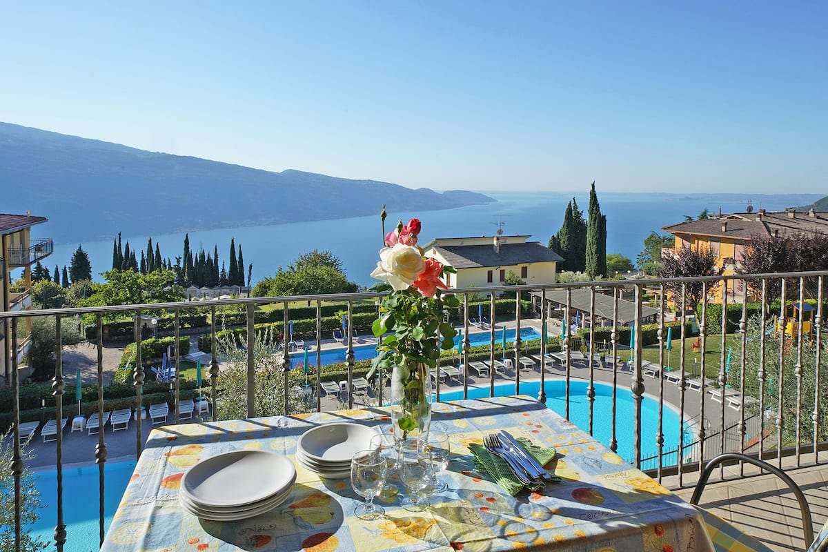 A terrace with a dining table set for four, overlooking a swimming pool and a mountainous landscape.