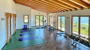 A spacious indoor gym with wooden beams, exercise equipment, and large windows offering a view of greenery. - Residence Besàss (Tignale)