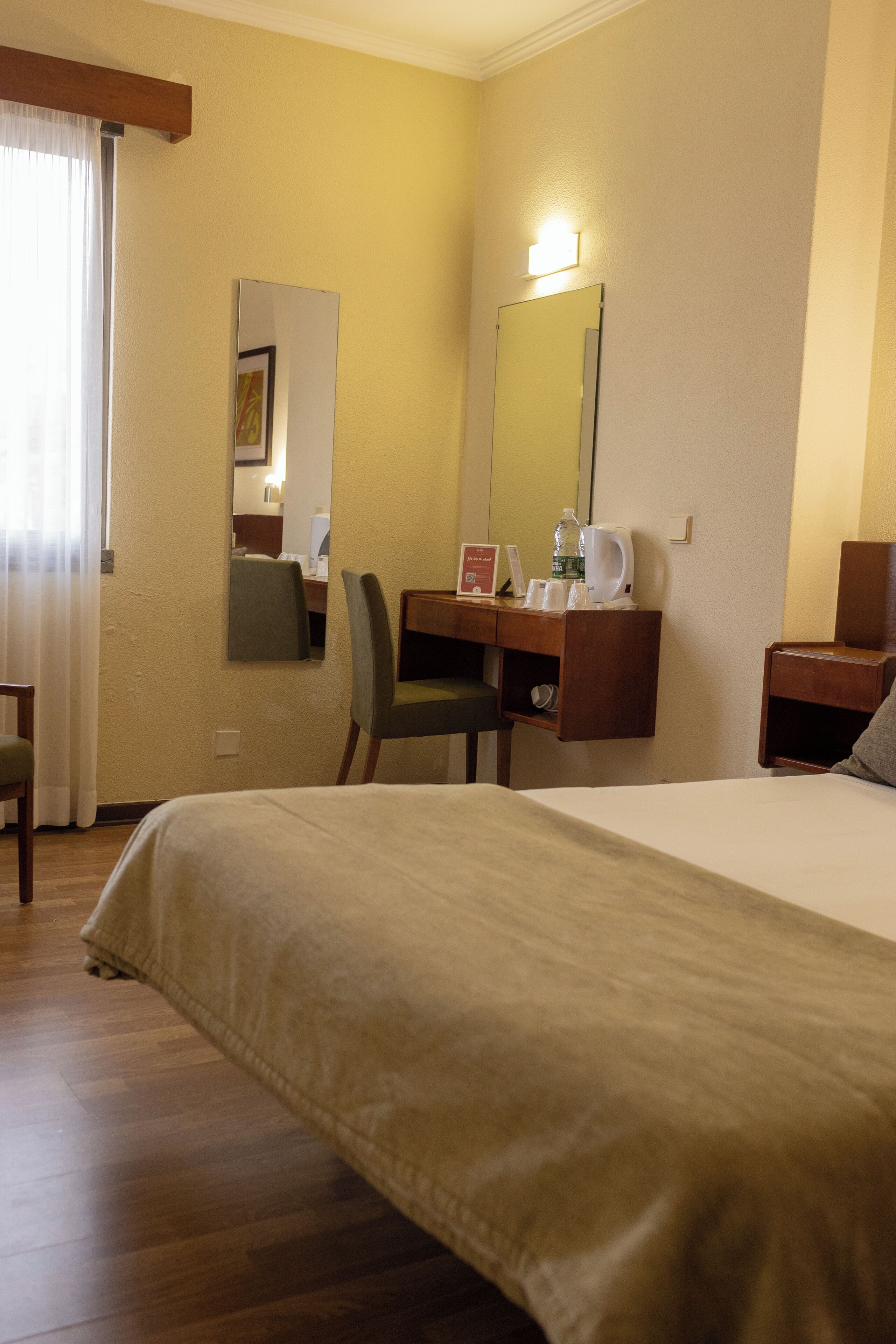 A hotel room with a bed, a desk with a chair, a mirror, and a nightstand.