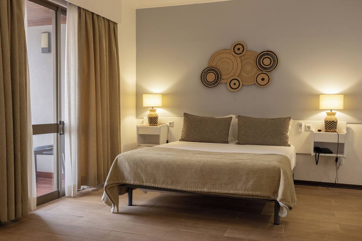 Ein Hotelzimmer mit einem Bett, zwei Nachttischen mit Lampen, einem Wandbild und einer Schiebetür zum Balkon.