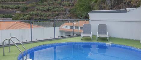 A rooftop swimming pool with two white lounge chairs, a metal ladder, and a view of a hillside with buildings.
