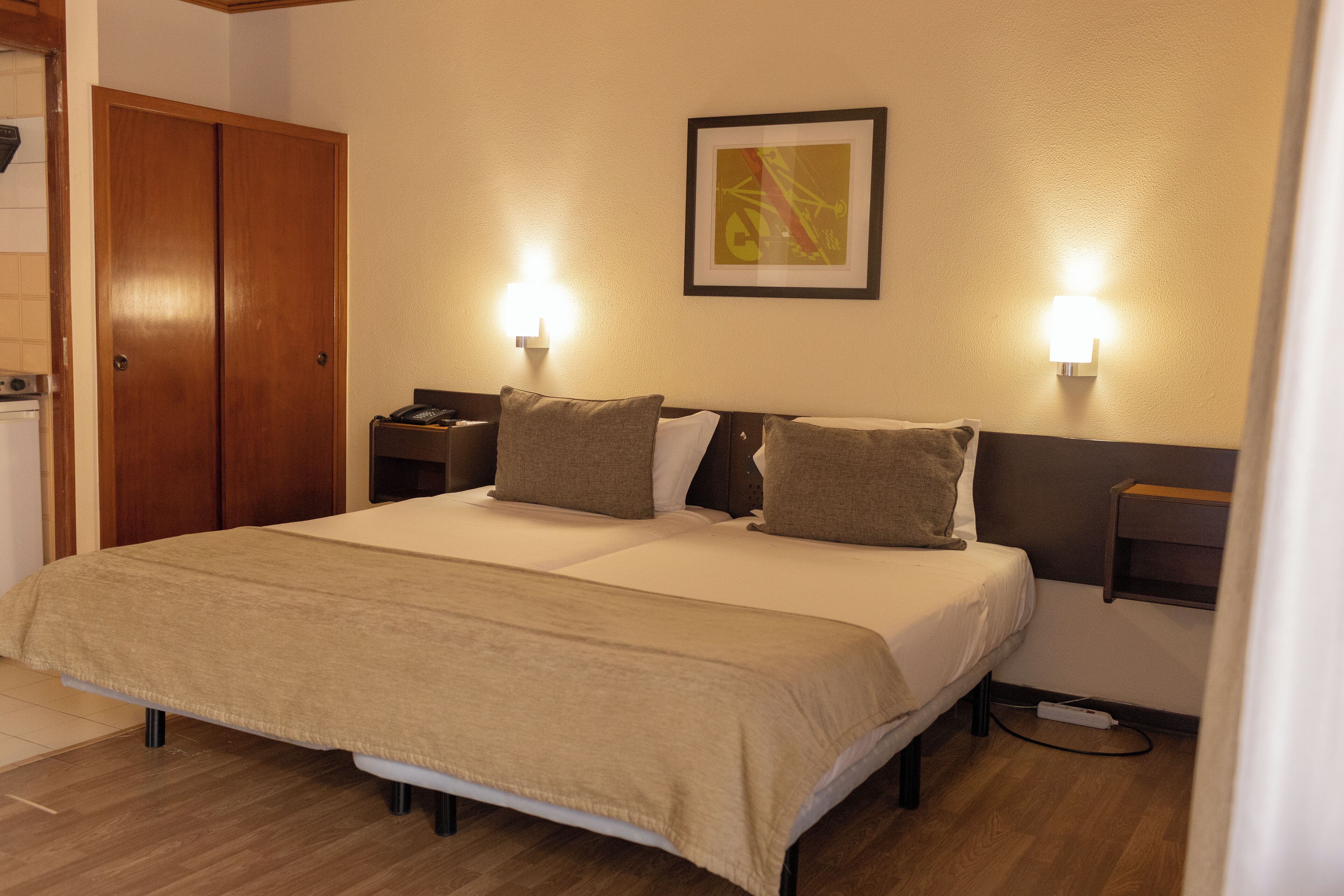 A hotel room with a double bed, two bedside tables, a nightstand, a wall-mounted lamp, and a framed picture on the wall.