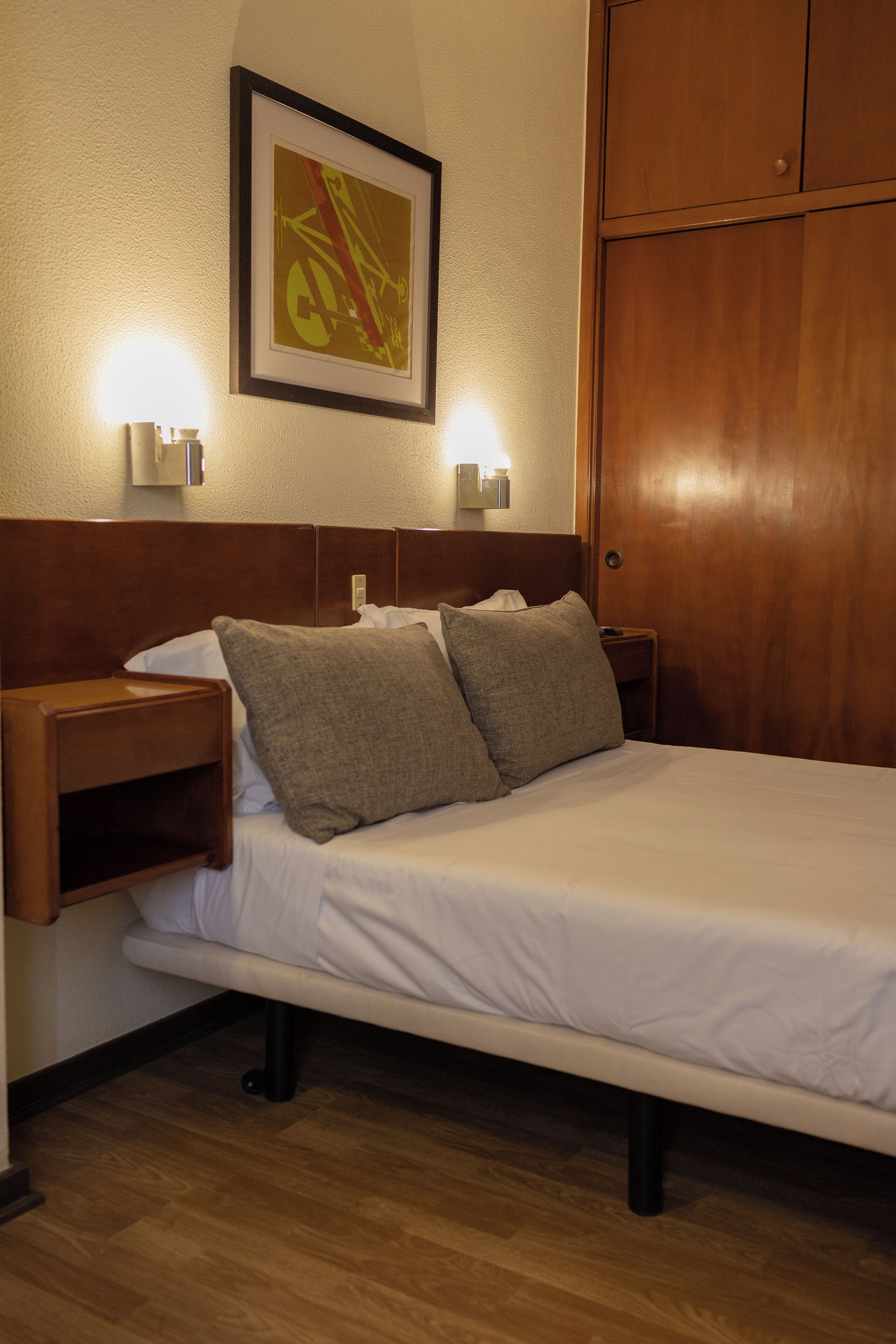 A hotel room with a single bed, two bedside lamps, a framed picture on the wall, and a wooden wardrobe.