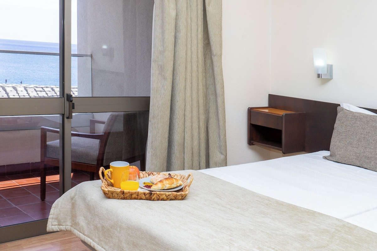 A hotel room with a bed, a bedside table, a window with a view of the sea, and a breakfast tray on the bed.