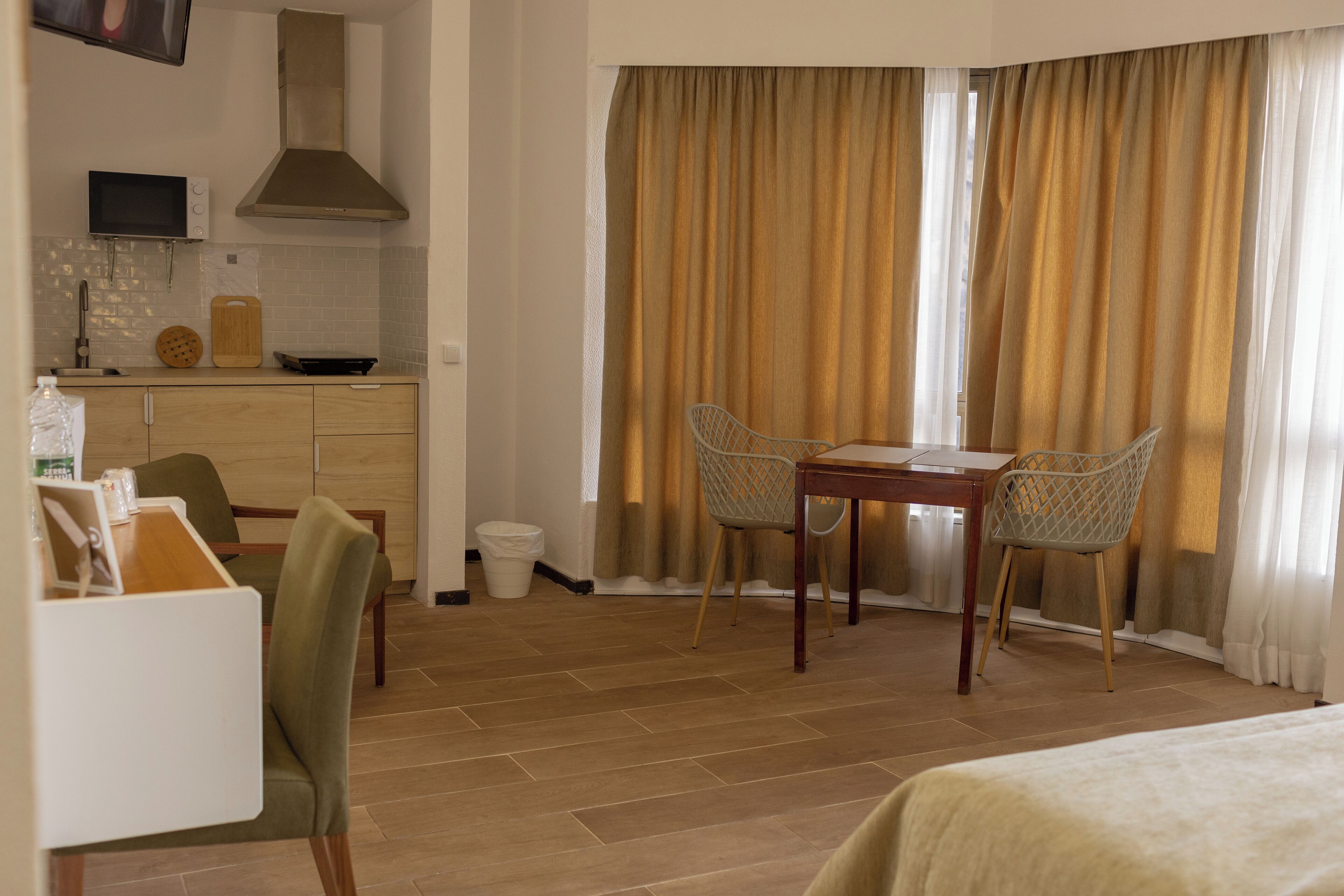 A hotel room with a wooden floor, a bed, a dining table with chairs, a kitchen area with a microwave, a cutting board, and a sink.