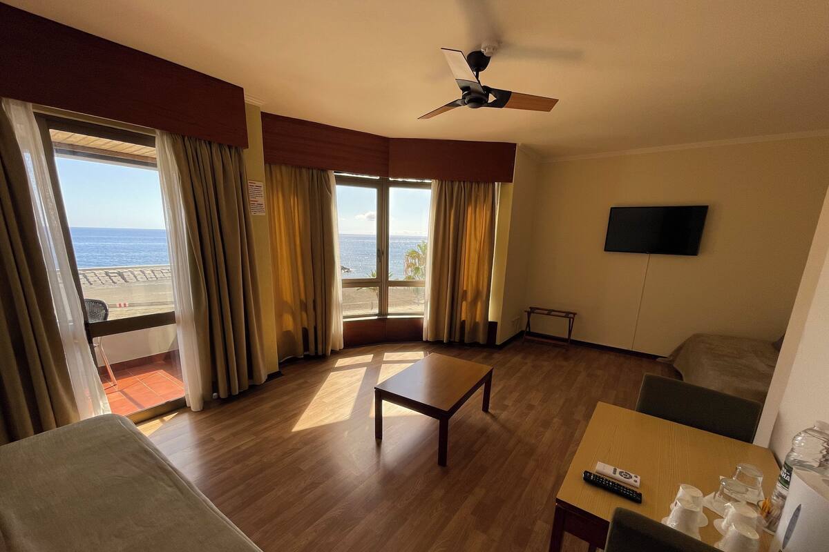 Ein Hotelzimmer mit Holzboden, einem Bett, einem Sofa, einem Tisch, einem Fernseher und einem Balkon mit Vorhängen.