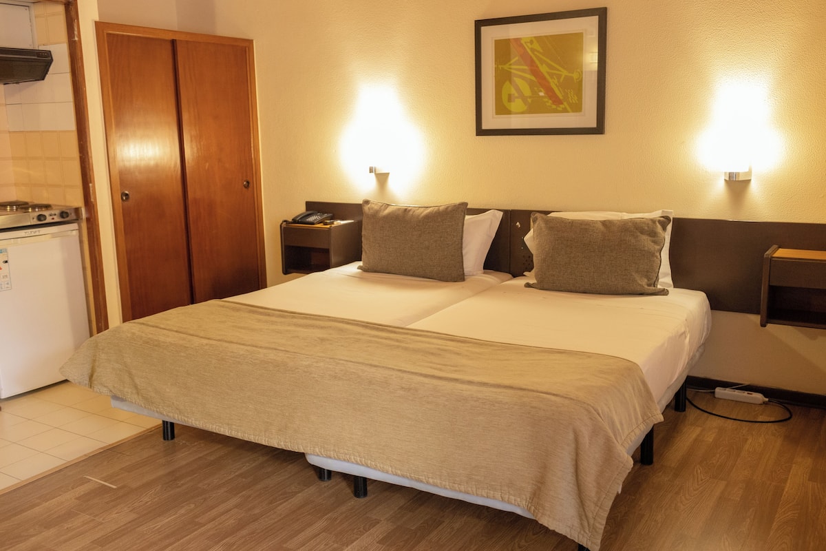 Ein Hotelzimmer mit Doppelbett, Nachttischen, einem kleinen Tisch, einem Kühlschrank und einem Holzschrank.