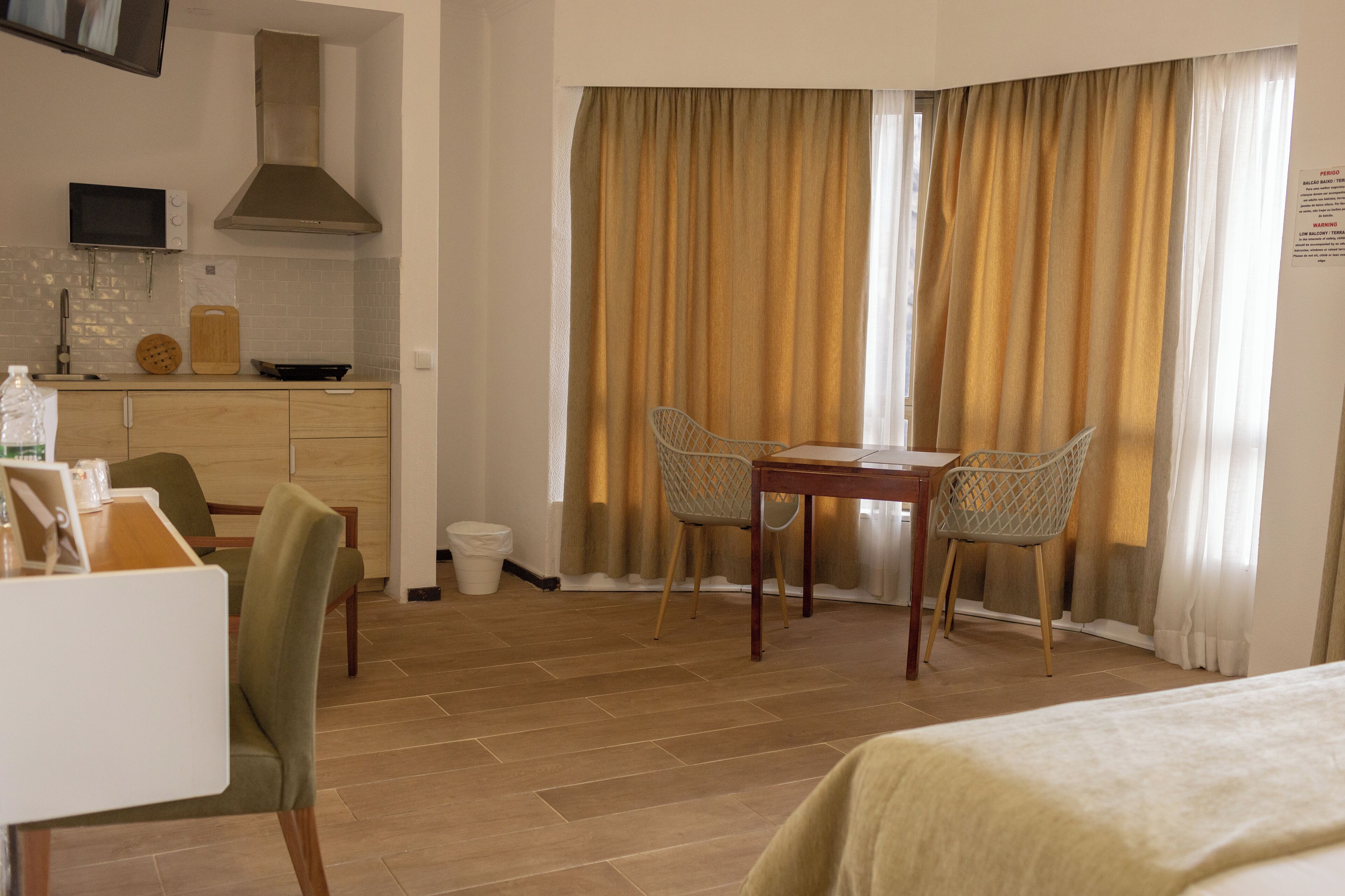A hotel room with a dining table, two chairs, a bed, a kitchen area with a microwave, and a window with curtains.