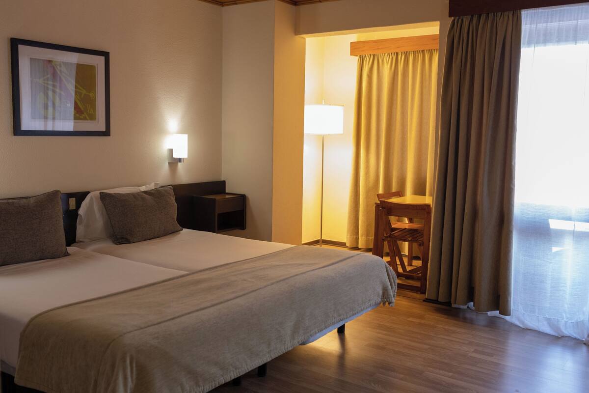 Ein Hotelzimmer mit zwei Betten, einem Nachttisch, einer Lampe, einem Sessel und einem Fenster mit Vorhängen.