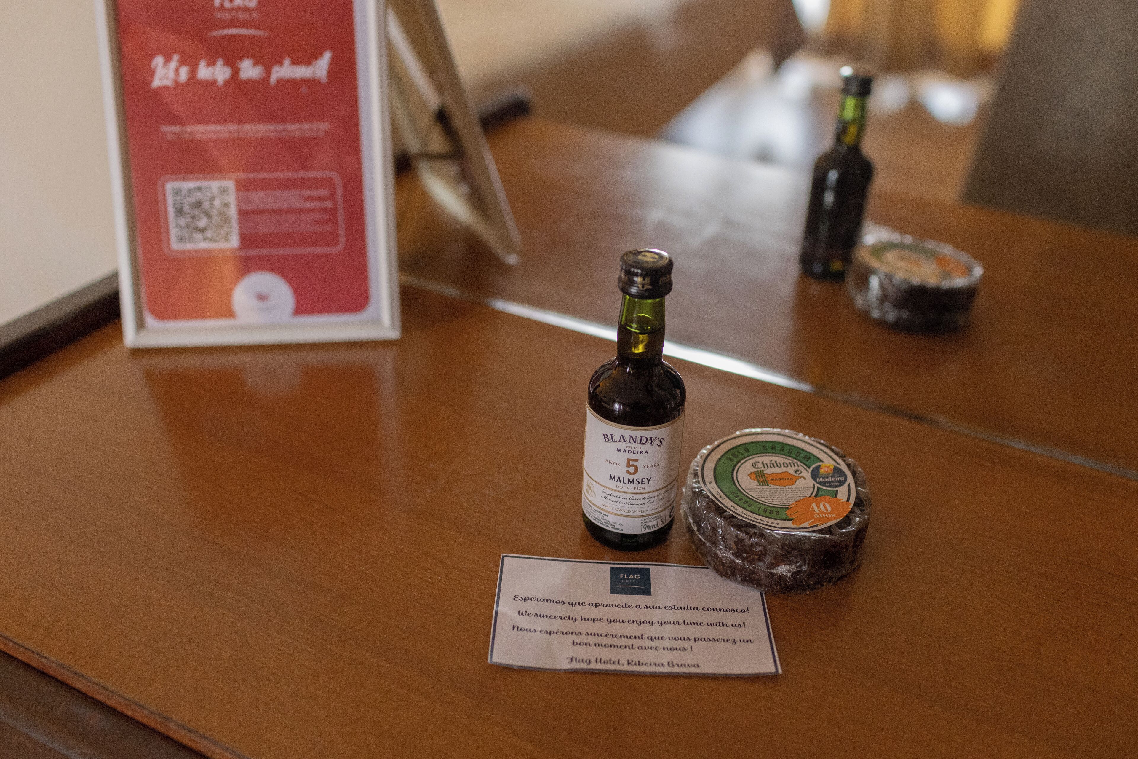 Una mesa de madera con una botella de Madeira Blandy, un frasco de mermelada y un cartel con un código QR.