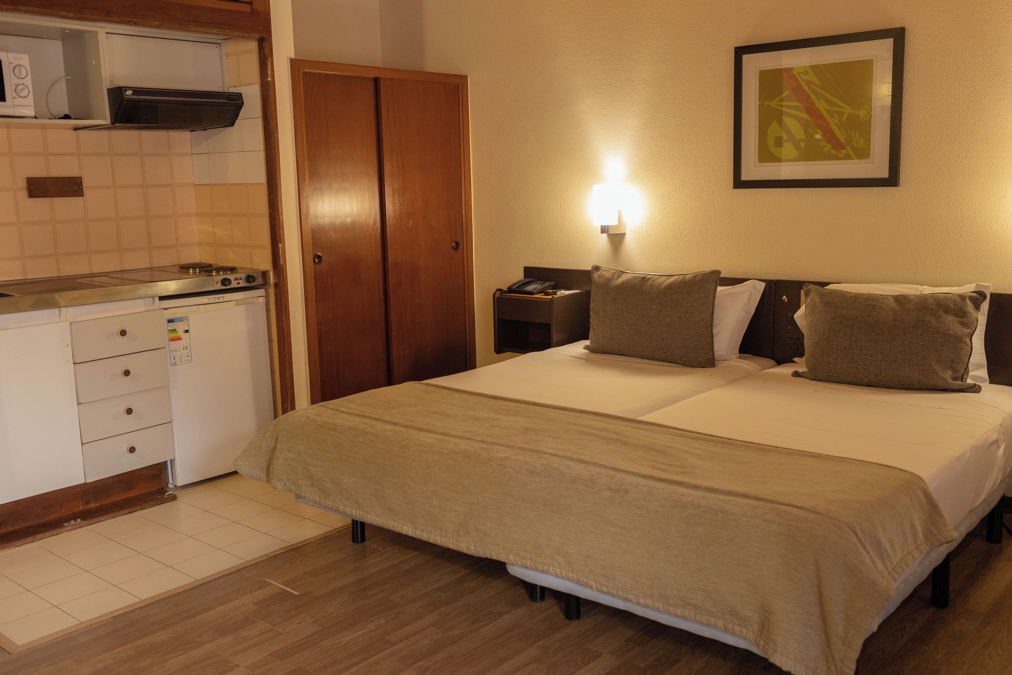 Ein Hotelzimmer mit Bett, Nachttisch, Lampe, Kleiderschrank und einer Küchenzeile mit Mikrowelle und kleinem Kühlschrank.