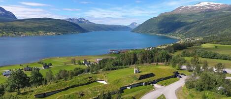 A panoramic view of a lakeside property with a winding road, green fields, and a small cluster of buildings.